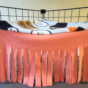 Peut inclure: Un hamac pour animaux de compagnie en polaire dans une cage. La couche supérieure présente un motif de formes noires, marron et orange sur fond blanc. La couche inférieure est pêche avec des franges verticales.