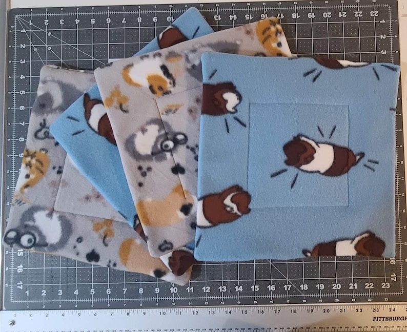May include: A set of four square fleece pads featuring a repeating pattern of brown, white, and black guinea pigs. The pads are layered, with one light blue pad on top, followed by two gray pads and one additional blue pad, all with stitched borders.