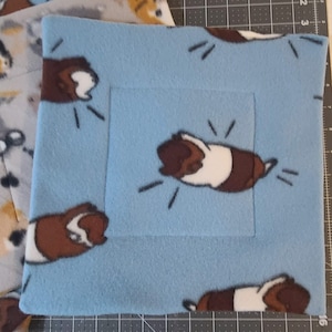 May include: A square light blue fleece fabric pad featuring a repeating pattern of brown and white guinea pigs. The fabric has a soft texture and is displayed on a cutting mat with visible inch markings along the edges for scale reference.