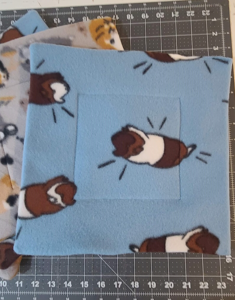 May include: A square light blue fleece fabric pad featuring a repeating pattern of brown and white guinea pigs. The fabric has a soft texture and is displayed on a cutting mat with visible inch markings along the edges for scale reference.