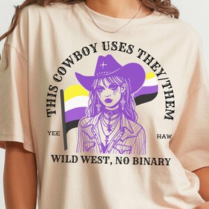 May include: Ivory-colored t-shirt featuring a graphic of a person in a cowboy hat with the text "This Cowboy Uses They/Them" and "Wild West, No Binary." The design includes the non-binary pride flag in purple, yellow, black, and white.