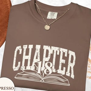 May include: Espresso-colored sweatshirt with the text "CHAPTER 18" in white, above an open book illustration. A gold-colored necklace with a round pendant rests on the shirt. The shirt is a Comfort Colors brand.