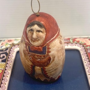 May include: A hand-painted, egg-shaped ornament depicting a woman in a red headscarf and traditional clothing. The ornament has a brass wire loop for hanging. The woman's face is detailed with a smiling expression. The ornament is on a blue tray.