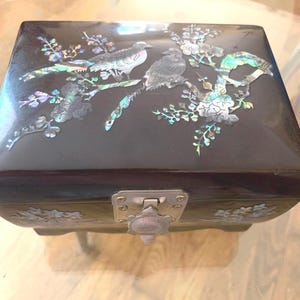 Può includere: Un portagioie nero con coperchio incernierato, con intarsi di uccelli e motivi floreali in madreperla. La scatola ha una chiusura in metallo e poggia su piedini. Gli elementi decorativi includono accenti iridescenti.