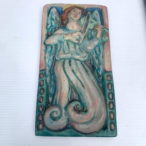 May include: A rectangular ceramic relief sculpture of an angel playing a stringed instrument. The angel has red hair, a yellow halo, and blue wings. The sculpture is painted in shades of blue, pink, and green.