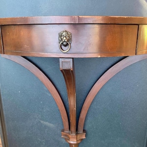 May include: A dark brown, semi-circular wall-mounted shelf with a drawer. The drawer features a brass lion's head pull. The shelf is supported by curved wooden brackets, creating a classic, elegant design. The shelf is approximately 30 inches wide.