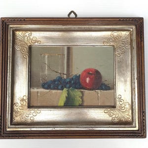 May include: A framed still life painting featuring a red apple, a bunch of dark grapes, and a glass. The painting is set against a neutral background and framed in a decorative gold and brown frame.