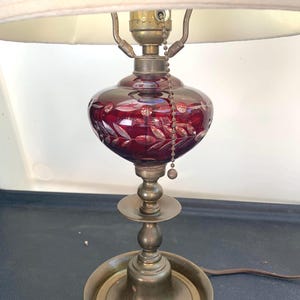 May include: A vintage table lamp with a ruby red glass base featuring a cut leaf design. The lamp has a brass-colored metal base and a cream-colored lampshade. A pull chain hangs from the lamp's top.