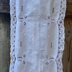 May include: White rectangular table runner with intricate floral embroidery and a delicate lace border. The runner features a repeating pattern of embroidered flowers and cut-out designs, adding a touch of elegance to any table setting.