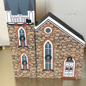 May include: A wooden church decoration with a stone-like facade. The church features arched windows adorned with wreaths and red bows. The steeple has a blue roof and white trim. A black cat sits on the roof.