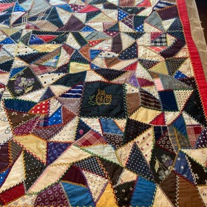 May include: A handmade quilt with a patchwork design. The quilt features a variety of fabrics in different colors and patterns, including triangles, squares, and other shapes. The quilt is bordered with a red fabric and has an embroidered owl design.