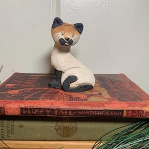 May include: A hand-carved cat figurine with a tan head and black ears, sitting on a stack of vintage books. The cat has a white body with black accents on its paws and tail. The books are red and green.