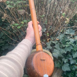 Op de afbeelding: Een handgemaakt houten snaarinstrument met een kokosnoot-resonator. Het instrument heeft een lange hals met stemknoppen en een kam. Het hout heeft een warme bruine kleur, tegen een achtergrond van groen gebladerte.