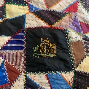 May include: A patchwork quilt constructed from various fabric scraps in diverse colors and patterns, including polka dots, stripes, and plaids. The pieces are joined by decorative cross-stitching. A central black square features yellow embroidered owls.