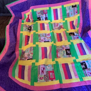 May include: A colorful quilt with a purple border and a patchwork design featuring cats. The quilt is made up of various shades of pink, yellow, green, and purple.