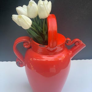 May include: A vibrant red ceramic pitcher with a handle and spout, holding a bouquet of white tulips. The pitcher has a glossy finish and decorative swirls. A striking floral arrangement for home decor.