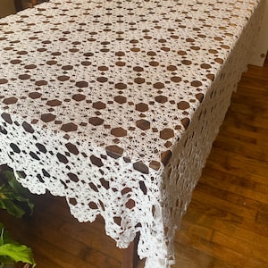 May include: A white, open-weave crochet tablecloth covers a wooden table. The tablecloth has a repeating pattern of geometric shapes and a scalloped edge. The table is rectangular and has a dark wood finish. The floor is hardwood.