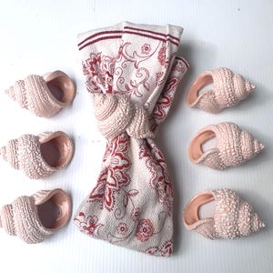 Könnte beinhalten: Ein Set aus sechs Muschel-Serviettenringen und einer gefalteten Serviette. Die Serviette ist weiß mit einem roten Blumenmuster. Die Serviettenringe sind hellrosa und in Form von Muscheln gehalten.