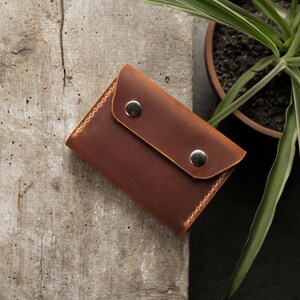 Closed bifold wallet in rectangular form showing camel crazy horse leather exterior with two brass snap closures. Compact dimensions approximately 4.5 by 3.35 inches. Matte waxy surface finish with subtle natural grain variation across the leather face.