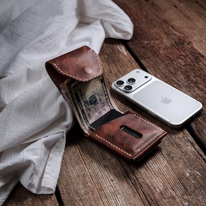 Pode incluir: Uma carteira de couro castanho com costuras visíveis, contendo dinheiro e um cartão preto. Um smartphone prateado está ao lado da carteira numa superfície de madeira. A carteira está aberta.