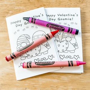 Puede incluir: Dos cuadernos blancos para colorear de San Valentín con el texto "Happy Valentine's Day Gnomie!" e ilustraciones de corazones. Tres crayones Crayola rosas y morados encima, listos para colorear.