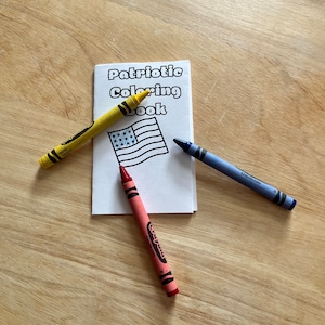 Puede incluir: Un pequeño libro para colorear con el título "Patriotic Coloring Book" y un contorno en blanco y negro de una bandera estadounidense. Tres lápices de colores están sobre una superficie de madera: amarillo, rojo y azul.