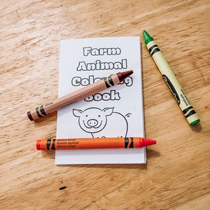 May include: A children's "Farm Animal Coloring Book" with a pig illustration, alongside three crayons: brown, green, and yellow-orange. The crayons are positioned around the book on a wooden surface.