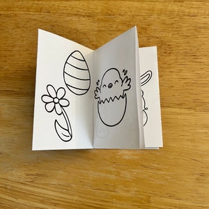 Puede incluir: Un pequeño folleto de papel blanco plegado con dibujos lineales negros con temática de Pascua. Las ilustraciones incluyen un huevo a rayas, una flor, un pollito saliendo de un huevo y un conejito. El folleto está abierto, mostrando varias páginas.