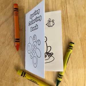 Puede incluir: Un pequeño "Libro para colorear de primavera" abierto, con dibujos lineales negros de una mariposa, una abeja y una rana. Dos crayones amarillos y uno naranja rodean el libro sobre una superficie de madera.