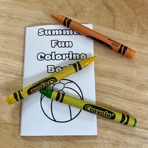 Könnte beinhalten: Ein weißes Malbuch mit dem Text "Summer Fun Coloring Book" und einer Strandball-Illustration. Drei Crayola-Buntstifte in Orange, Gelb und Grün liegen um das Buch herum.