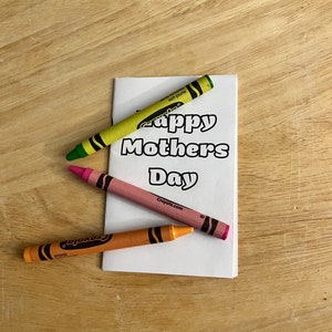 Può includere: Un biglietto bianco con la scritta "Happy Mothers Day" è circondato da tre pastelli gialli, rosa e arancioni. I pastelli sono su una superficie di legno marrone chiaro.