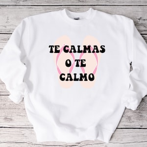 以下が含まれることがあります： 白いスウェットシャツに、黒い文字で「Te Calmas O Te Calmo」と書いてあり、ピンクのビーチサンダルが描かれています。