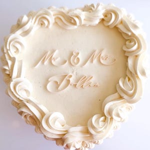 Op de afbeelding: Een hartvormige taart met witte glazuur en decoratieve krullen langs de rand. De taart is voorzien van de woorden "Mr & Mrs Bellia" in elegant schrift, perfect voor een bruiloft of jubileum.