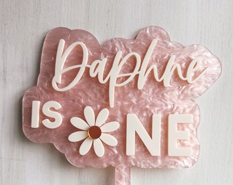 May include: Pink and white cake topper with the text "Daphne is one" and a white daisy with a gold center.