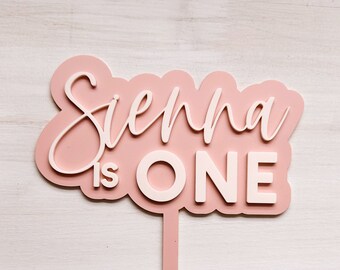 May include: Pink cake topper with the text "Sienna is ONE" in white lettering. The topper is on a pink stick.