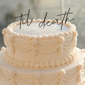 May include: A two-tiered wedding cake with cream-colored frosting and decorative piping. The cake is adorned with the phrase "til death" in a delicate black script cake topper. The cake has a classic, elegant design.