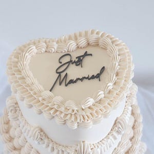 Op de afbeelding: Een bruidstaart met drie lagen en een hartvormige bovenkant. De taart is versierd met witte frosting en ingewikkelde spuitdetails. De bovenste laag heeft de woorden "Just Married" in zwart.