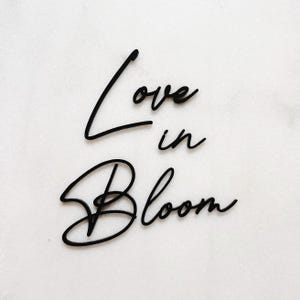 Może przedstawiać: Czarne litery w stylu pisma odręcznego na białym tle, które brzmią "Love in Bloom".