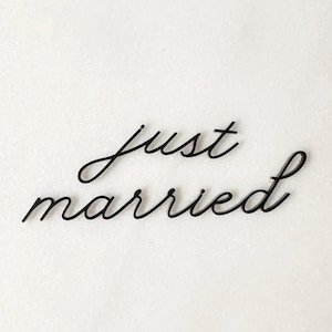 Op de afbeelding: Zwarte cursieve letters die "just married" spellen op een witte achtergrond. De elegante belettering is een decoratief element, perfect voor bruiloftsdecoraties of aankondigingen. De woorden zijn in een vloeiende, handgeschreven stijl.