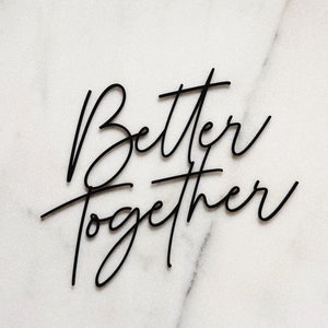 Op de afbeelding: Zwarte cursieve tekst op een witte marmeren achtergrond leest "Better Together". De woorden zijn gestapeld, met "Better" boven "Together". De belettering is elegant en modern, geschikt voor woondecoratie of bruiloftsborden.
