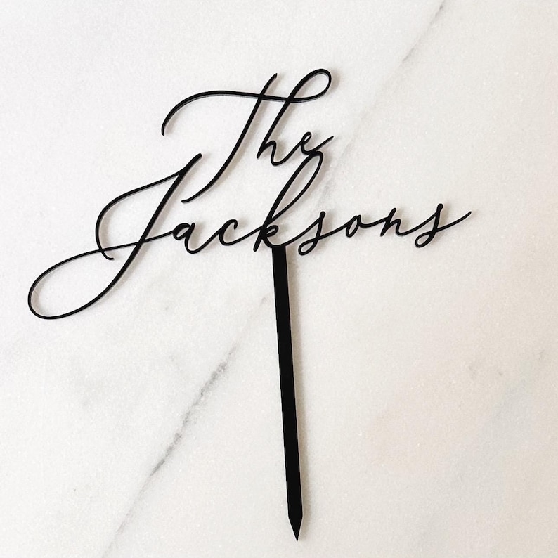 K&ouml;nnte beinhalten: Schwarzer Tortenaufsatz mit den Worten "The Jacksons" in eleganter Schreibschrift. Der Aufsatz hat einen langen, spitzen Stiel zum Einsetzen in einen Kuchen. Der Hintergrund ist eine wei&szlig;e Marmoroberfl&auml;che.