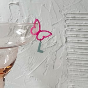 Pode incluir: Um encanto de bebida em forma de borboleta rosa com um pequeno pingente de letra "L" azul claro, preso à borda de um copo tingido de rosa. O fundo apresenta uma parede branca texturizada.
