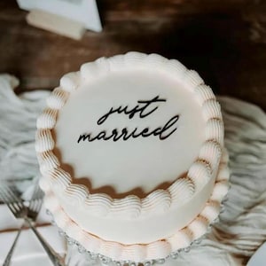 Op de afbeelding: Een ronde bruidstaart met twee lagen, witte glazuur en een decoratieve rand. De bovenkant van de taart is voorzien van de woorden "just married" in elegant zwart schrift. De taart staat op een heldere, kralen taartstandaard.