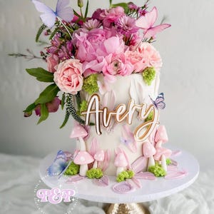 May include: A two-tiered white cake decorated with pink flowers, butterflies, and mushroom accents. The cake is topped with a floral arrangement of pink and purple flowers. The name "Avery" is written on the front of the cake in gold script. The cake sits on a white cake stand.