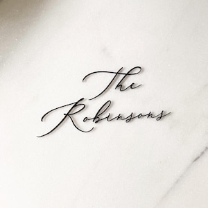 May include: Black cursive text on a white marble background reads "The Robinsons." The elegant script is a decorative element, likely for home decor or event signage. The overall aesthetic is minimalist and modern.