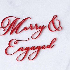 Op de afbeelding: Rode cursieve tekst die "Merry & Engaged" leest op een wit marmeren oppervlak. De woorden zijn in een vloeiend script, perfect voor bruiloft- of verlovingsaankondigingen en decoraties.
