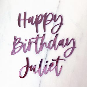 Op de afbeelding: Een magenta taarttopper met de woorden "Happy Birthday Juliet" in een sierlijk lettertype. Ontworpen om op een verjaardagstaart te plaatsen. De achtergrond is een wit marmeren oppervlak. Het reflecterende oppervlak voegt elegantie toe.