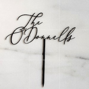K&ouml;nnte beinhalten: Schwarzer Tortenaufsatz mit den Worten "The O'Donnells" in eleganter Schreibschrift. Der Aufsatz hat einen langen, spitzen Stiel zum Einstecken in einen Kuchen. Der Hintergrund ist eine wei&szlig;e Marmoroberfl&auml;che.