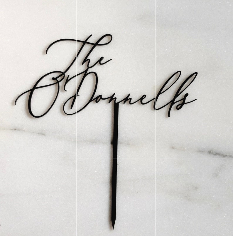 K&ouml;nnte beinhalten: Schwarzer Tortenaufsatz mit den Worten "The O'Donnells" in eleganter Schreibschrift. Der Aufsatz hat einen langen, spitzen Stiel zum Einstecken in einen Kuchen. Der Hintergrund ist eine wei&szlig;e Marmoroberfl&auml;che.