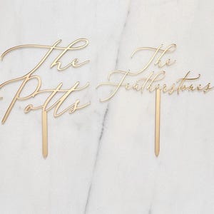 K&ouml;nnte beinhalten: Zwei goldfarbene Tortenaufs&auml;tze mit eleganter Schreibschrift. Einer tr&auml;gt die Aufschrift "The Potts" und der andere "The Featherstones". Jeder Aufsatz hat einen schmalen, spitzen Stab zum Einstecken in einen Kuchen.